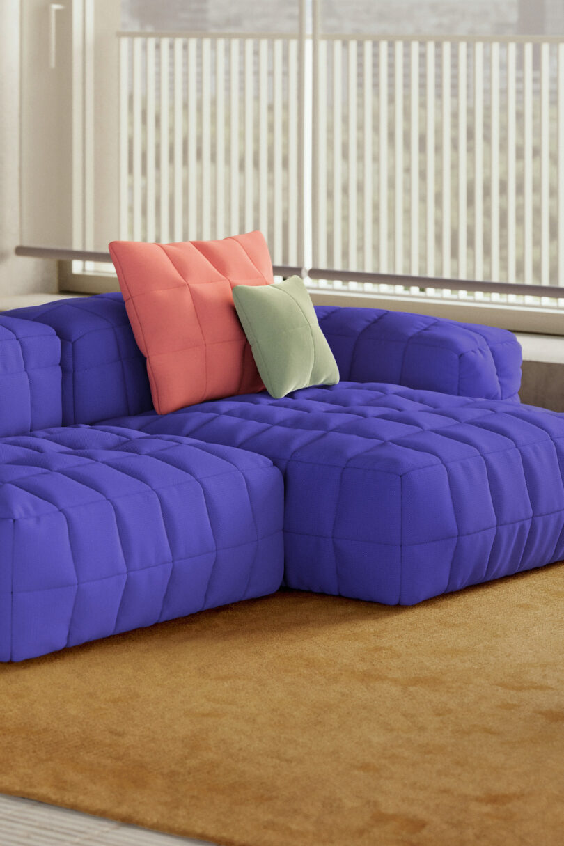 A modern blue sectional sofa with a tufted design, topped with one coral pillow and one light green pillow, sits on a tan carpet in front of large windows with vertical blinds