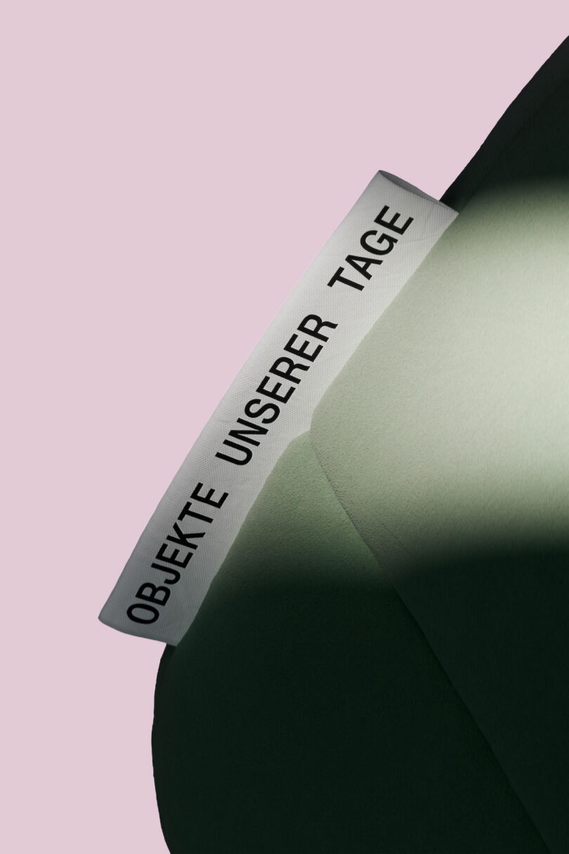 A white fabric label with the text "OBJEKTE UNSERER TAGE" is attached to a green fabric surface, against a pale pink background