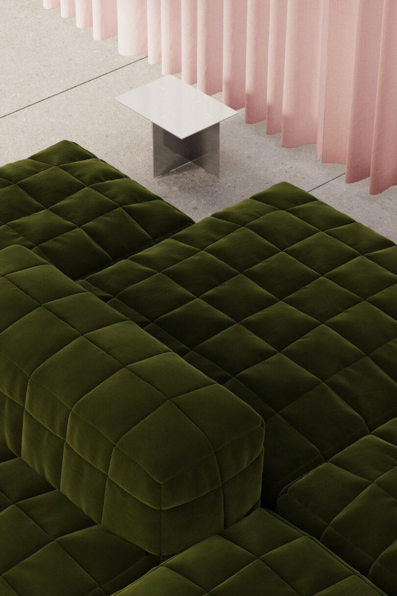 Green velvet tufted sectional sofa with square pattern, next to a small white modern side table and pink sheer curtains on a concrete floor