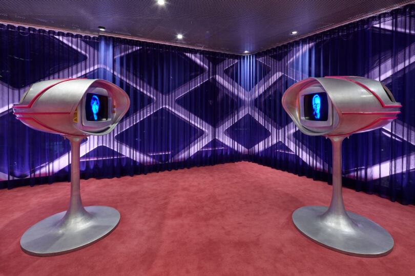 Two futuristic silver kiosks with screens displaying faces stand on a red carpeted floor against a blue and purple geometric backdrop.