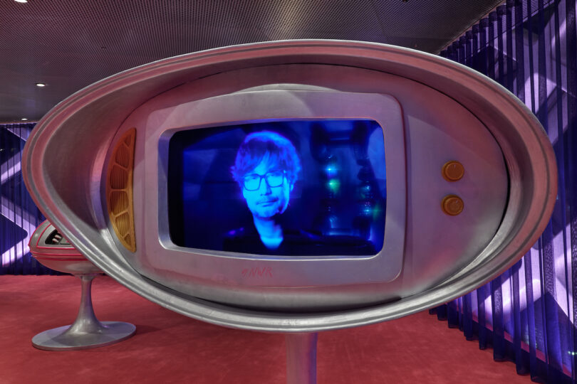 A retro-style television displays a blue-tinted image of a person’s face, with purple lighting and draped curtains in the background.