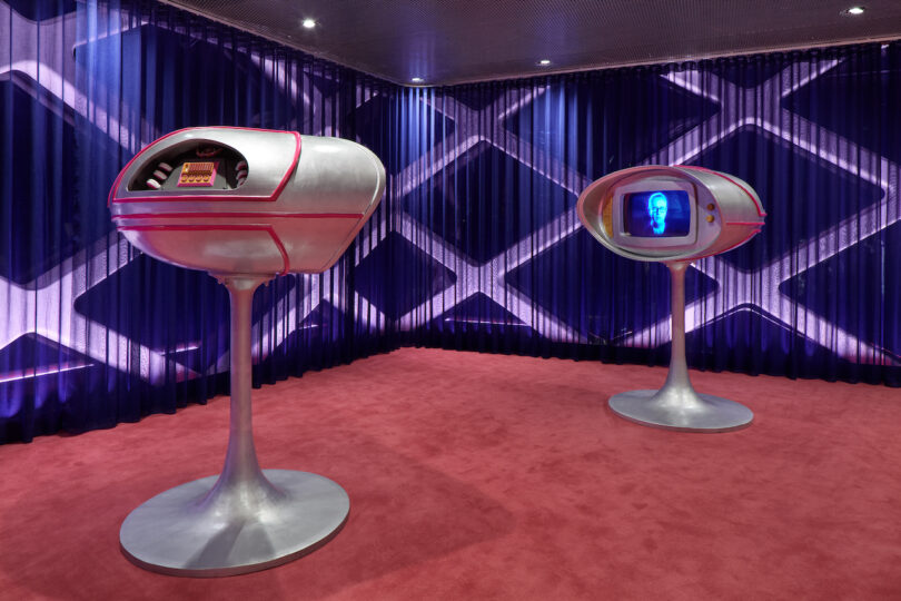 Two futuristic silver kiosks with screens stand on a red carpet in a room with blue geometric-patterned walls and ceiling spotlights