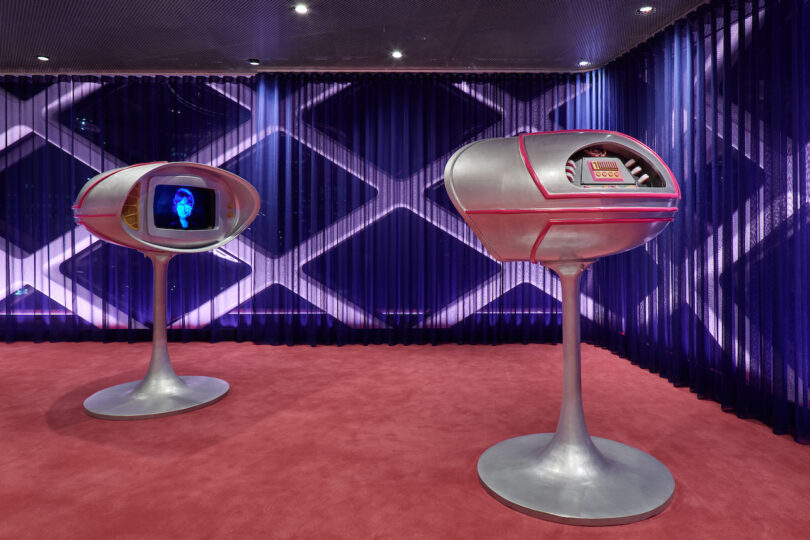 Two futuristic silver kiosks with screens stand on a red carpet in a room with blue geometric-patterned walls and ceiling spotlights