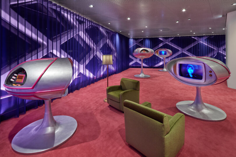 Room with futuristic silver pods on pedestals, each displaying a digital face on screens, pink carpet, green armchair, and geometric patterned curtains