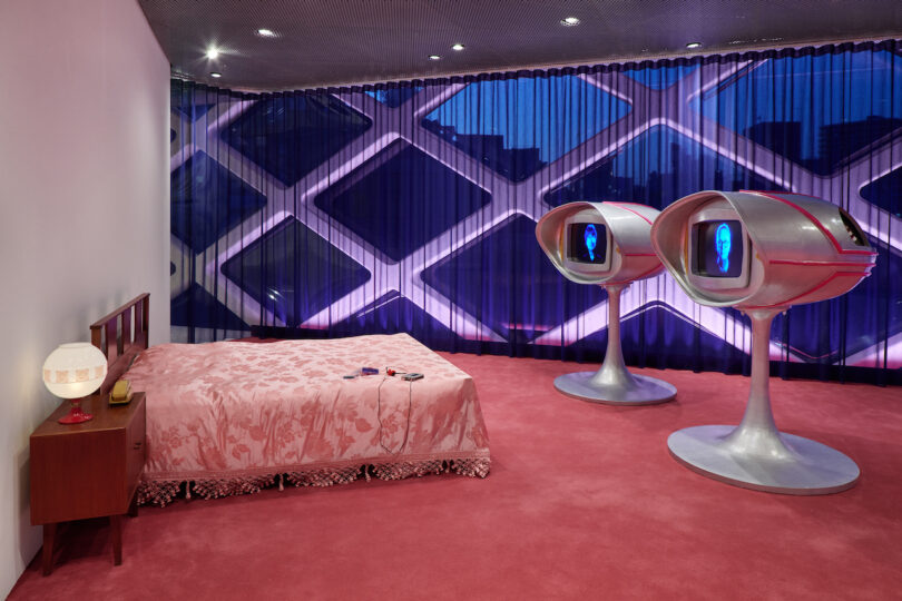 Retro-futuristic bedroom with pink carpet, a bed with a floral bedspread, a vintage nightstand, and two pod-shaped TV screens on stands in front of a geometric-patterned window