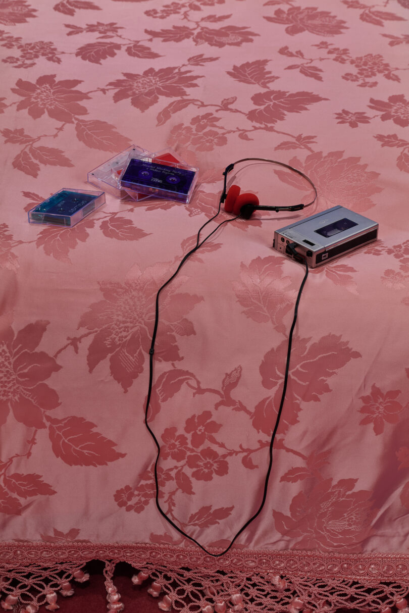 A portable cassette player with red headphones, two cassette tapes, and a clear case rest on a pink floral-patterned bedspread