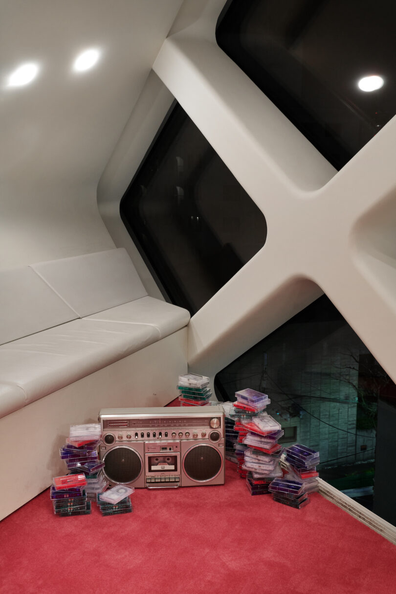 A silver boombox sits on a red carpet next to stacks of cassette tapes in a modern room with large angular windows and a white sofa