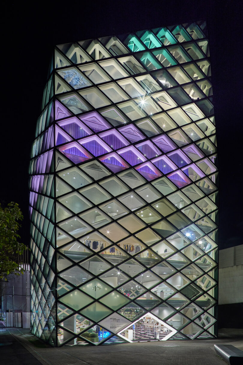 A multi-story glass building with a diamond-patterned facade is illuminated at night with interior and exterior lights, revealing several floors inside