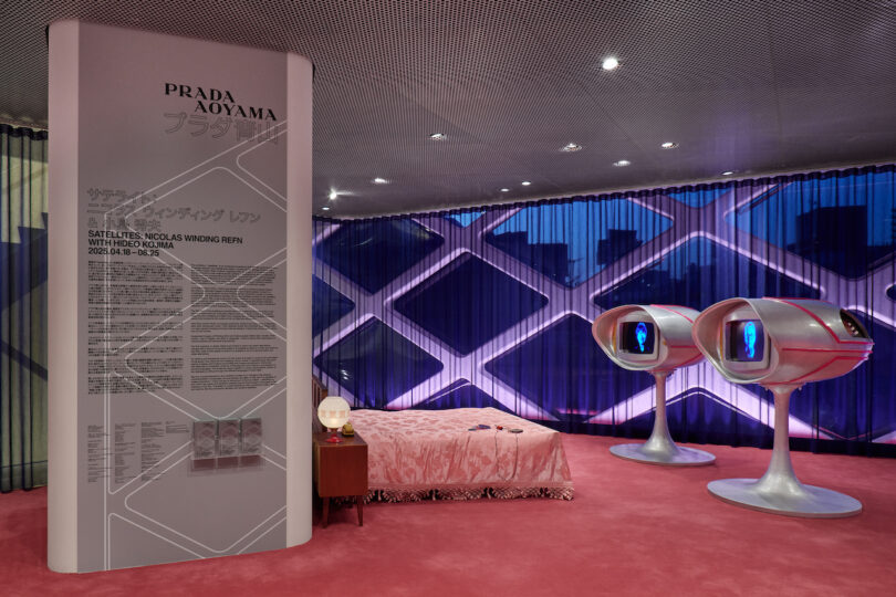 A modern exhibition room with a large display panel, a pink bed, a vintage nightstand, and two futuristic pod-like chairs against a purple geometric window backdrop