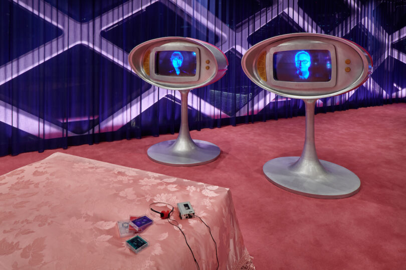 Two retro-futuristic television sets display a man's face in blue on screens; a bed with pink bedding and headphones rests in the foreground, all in a pink-carpeted room with patterned walls
