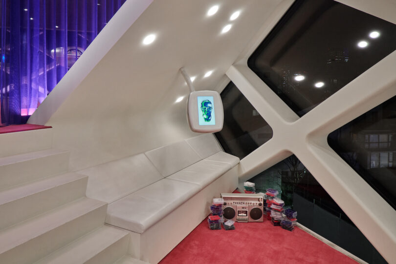 Modern room with white built-in seating, a wall-mounted screen, stacks of cassette tapes, a retro radio, and large geometric windows overlooking a nighttime cityscape