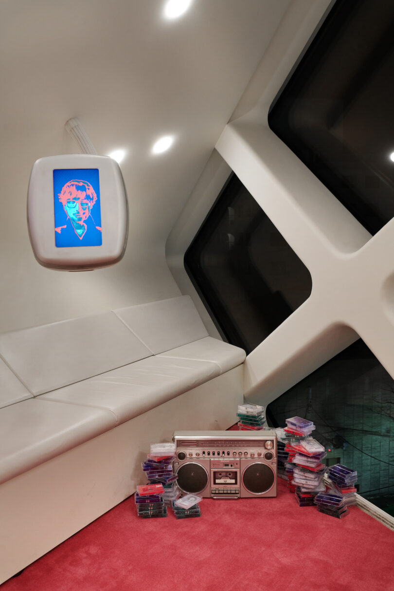 A modern white lounge with a curved sofa, a wall-mounted screen displaying pop art, a large boombox, and stacks of cassette tapes on a red carpet