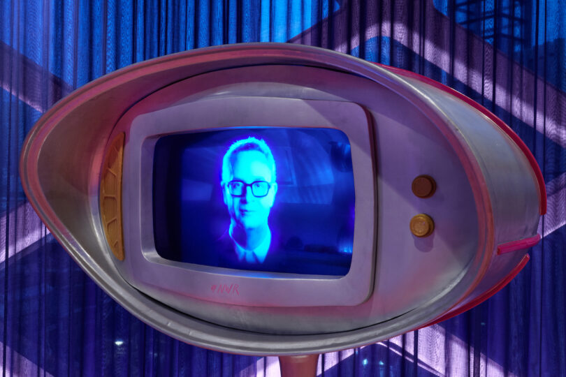 A retro-style television displays a blue-tinted image of a person’s face, with purple lighting and draped curtains in the background.
