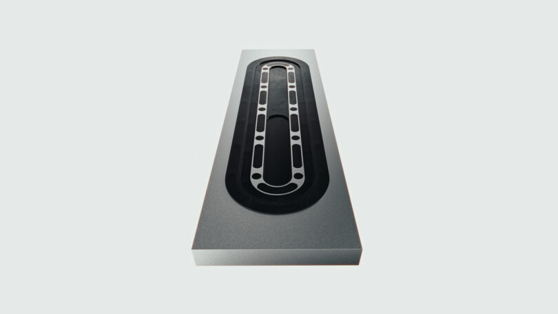 A rectangular metal block reminiscent of a cleaver, featuring an elongated oval cutout and multiple circular holes evenly spaced along the inner edge.