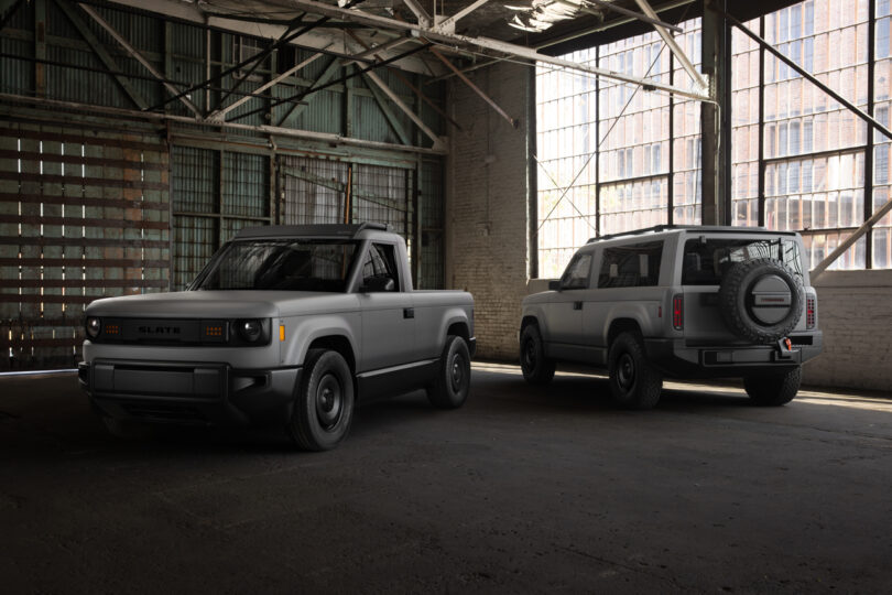 Two gray electric vehicles, a pickup truck and an SUV, are parked inside an industrial warehouse with large windows and exposed beams.