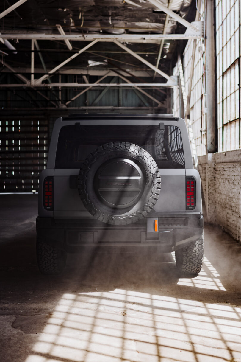 A grey SUV is parked inside an industrial warehouse, illuminated by sunlight streaming through large windows, with dust visible on the ground.
