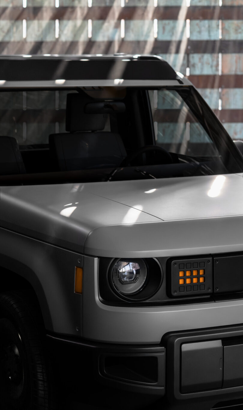 The front half of a modern, boxy silver SUV with a square grille and single round headlight, parked indoors with sunlight streaming through slats above.