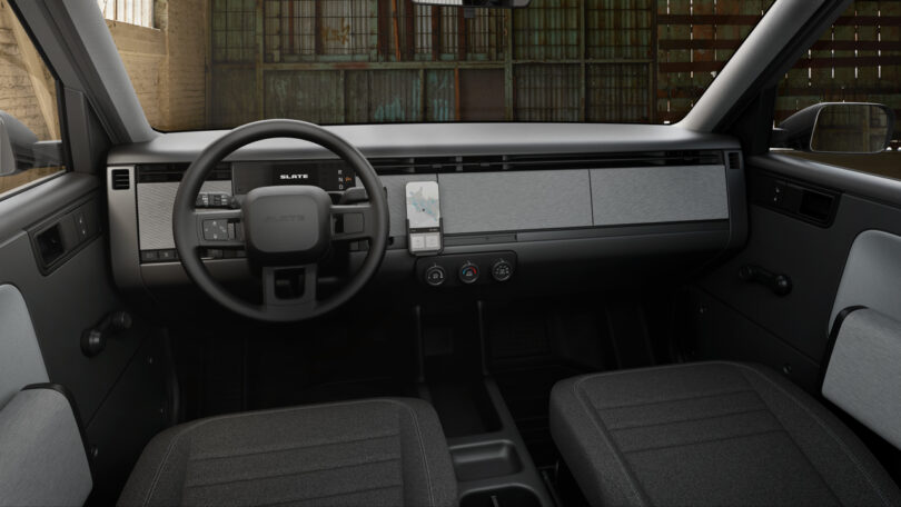 A minimalist car interior features a flat dashboard, wide touchscreen display, steering wheel, and two front seats, with a warehouse-like background visible through the windshield.