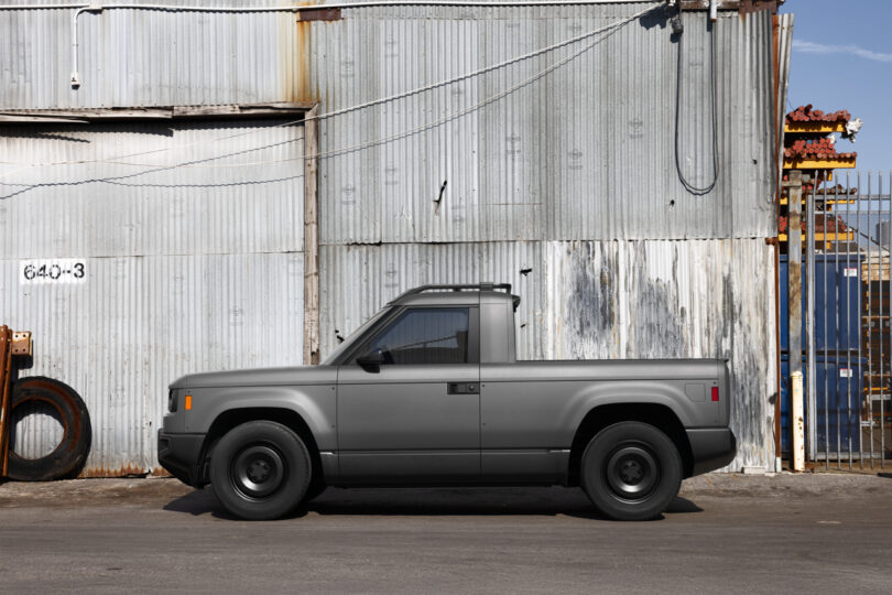 A matte grey compact pickup truck is parked in front of a corrugated metal industrial building.
