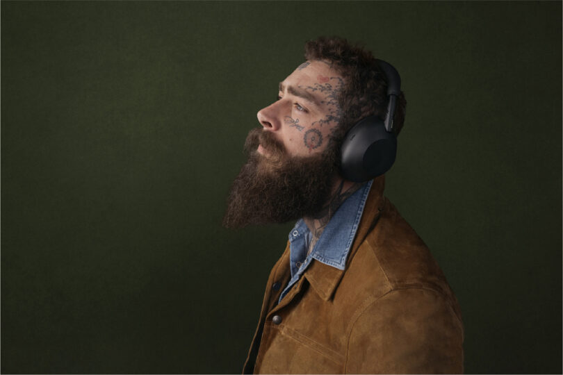A bearded man with facial tattoos wears large black WH-1000XM6 headphones and a brown suede jacket over a denim shirt, facing left against a plain dark green background.