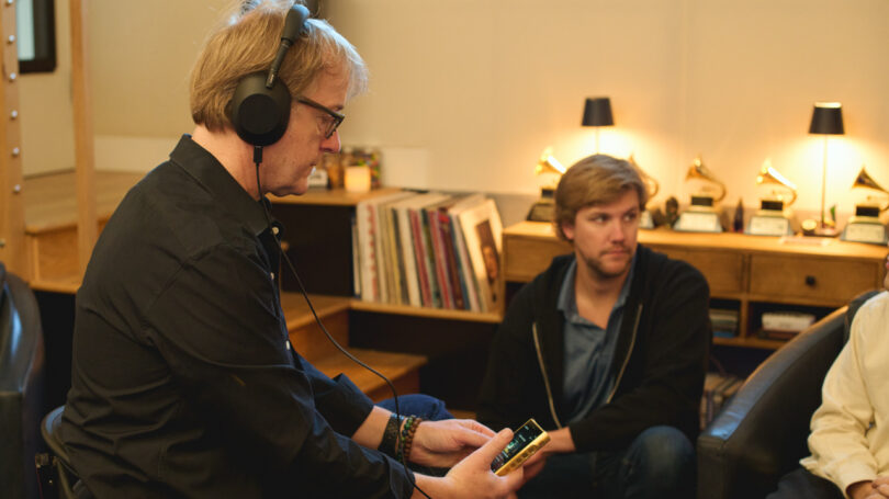 A man wearing WH-1000XM6 headphones holds a device, while another man sits nearby in a room filled with shelves of books, records, and Grammy awards.