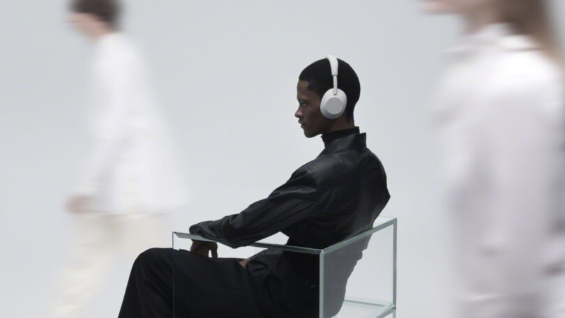 A person wearing WH-1000XM6 white headphones and a black outfit sits in a glass chair, while blurred figures in white move past in the background.