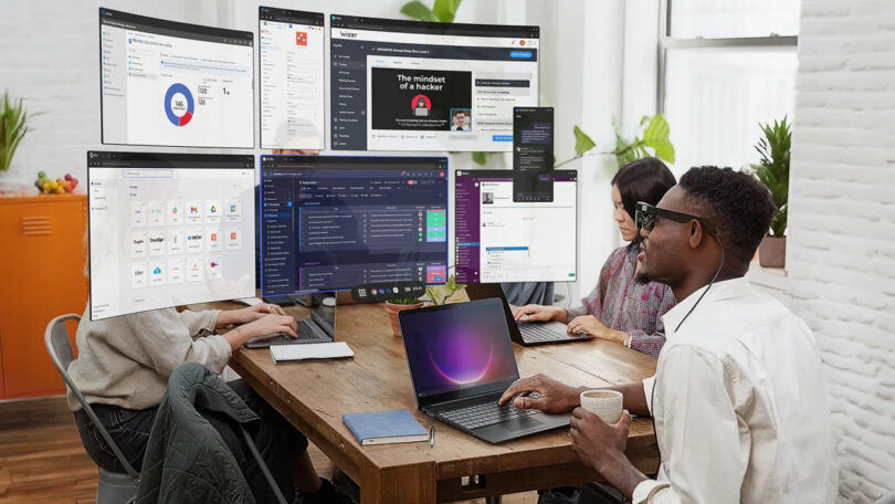Three people sit at a table using laptops, each wearing AR glasses that display multiple virtual computer screens around them.