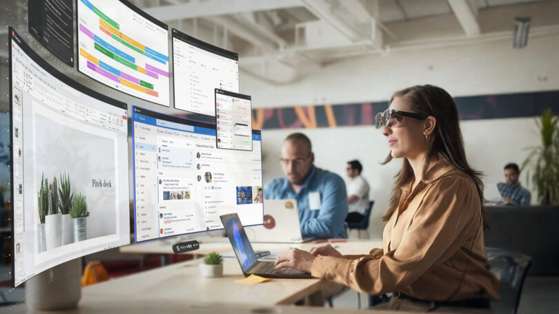 A woman wearing augmented reality glasses works on a laptop at a desk, viewing multiple virtual screens in a modern office setting.