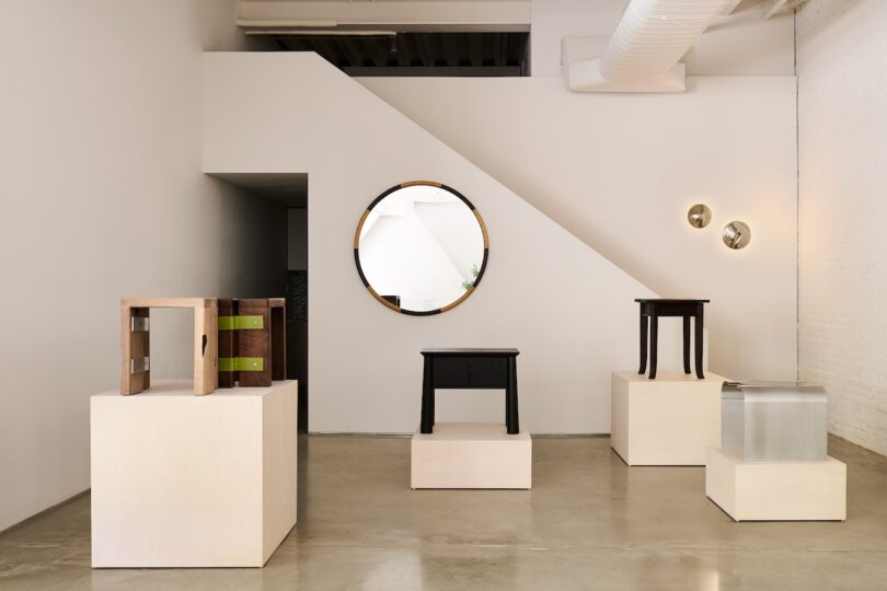 A minimalist gallery room displaying five unique side tables on white pedestals, with a round mirror and two wall lights on a white wall under a staircase.