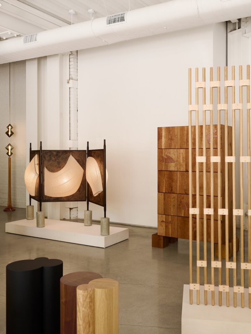 A modern interior space features sculptural wooden furniture, a geometric room divider, a large lamp with curved shades, and a tall floor lamp against a white wall.