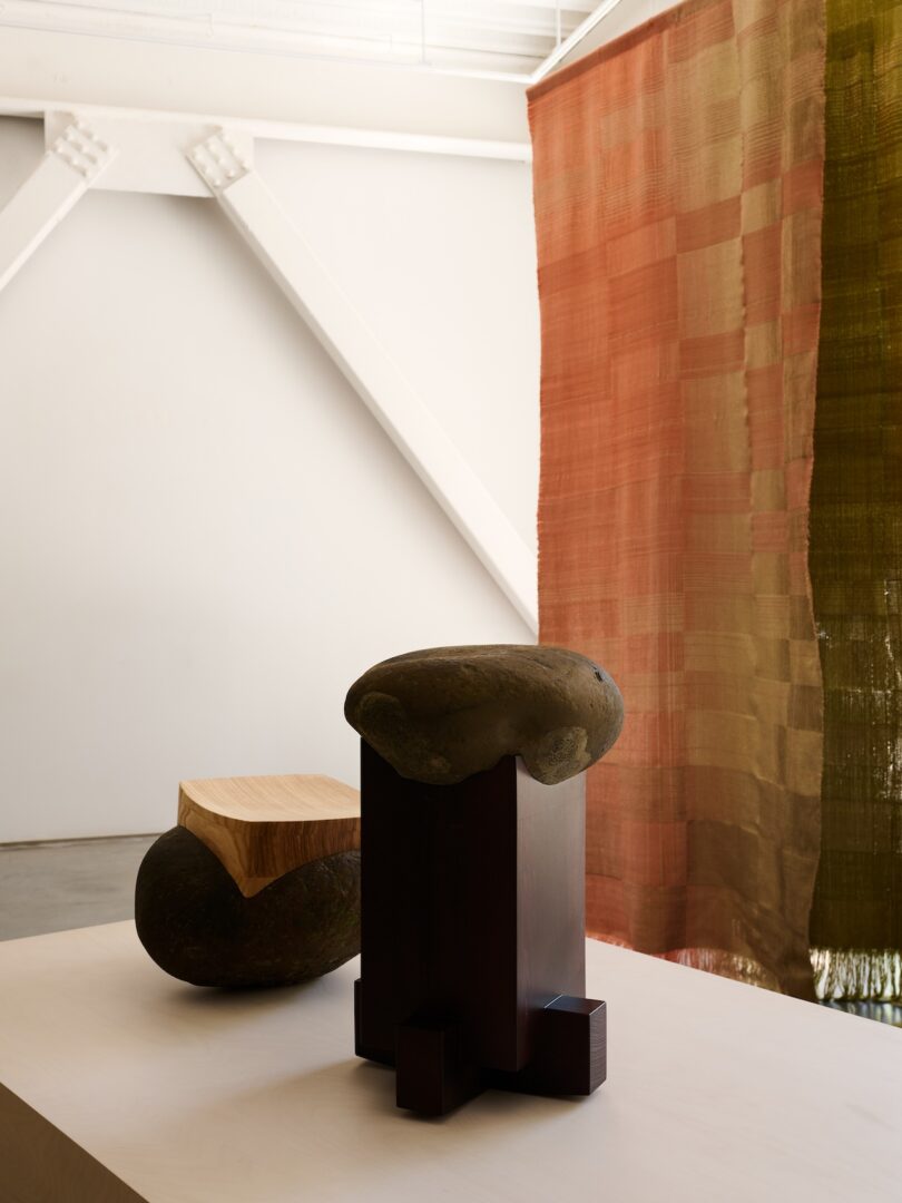 A minimalist interior with two abstract sculptures on a white table, positioned near a red and olive green woven curtain.