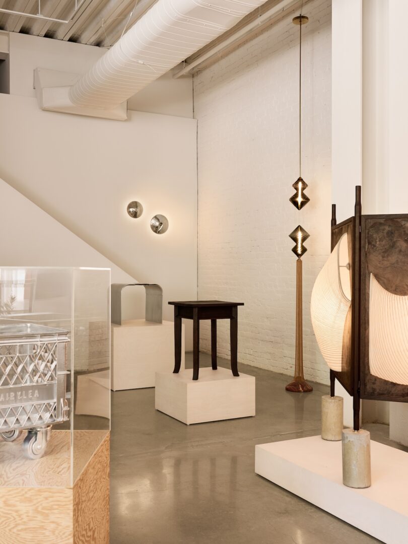 A modern gallery with minimalist furniture, sculptures, and lighting fixtures displayed on plinths against white brick walls and polished concrete floors.