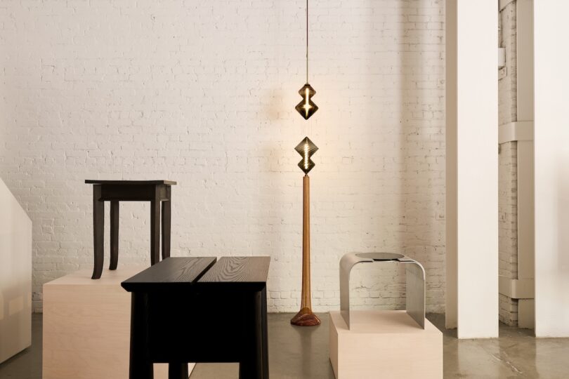 A minimalist room with light brick walls features modern furniture, including tables and a stool on wooden platforms, and a unique floor lamp with geometric shades.