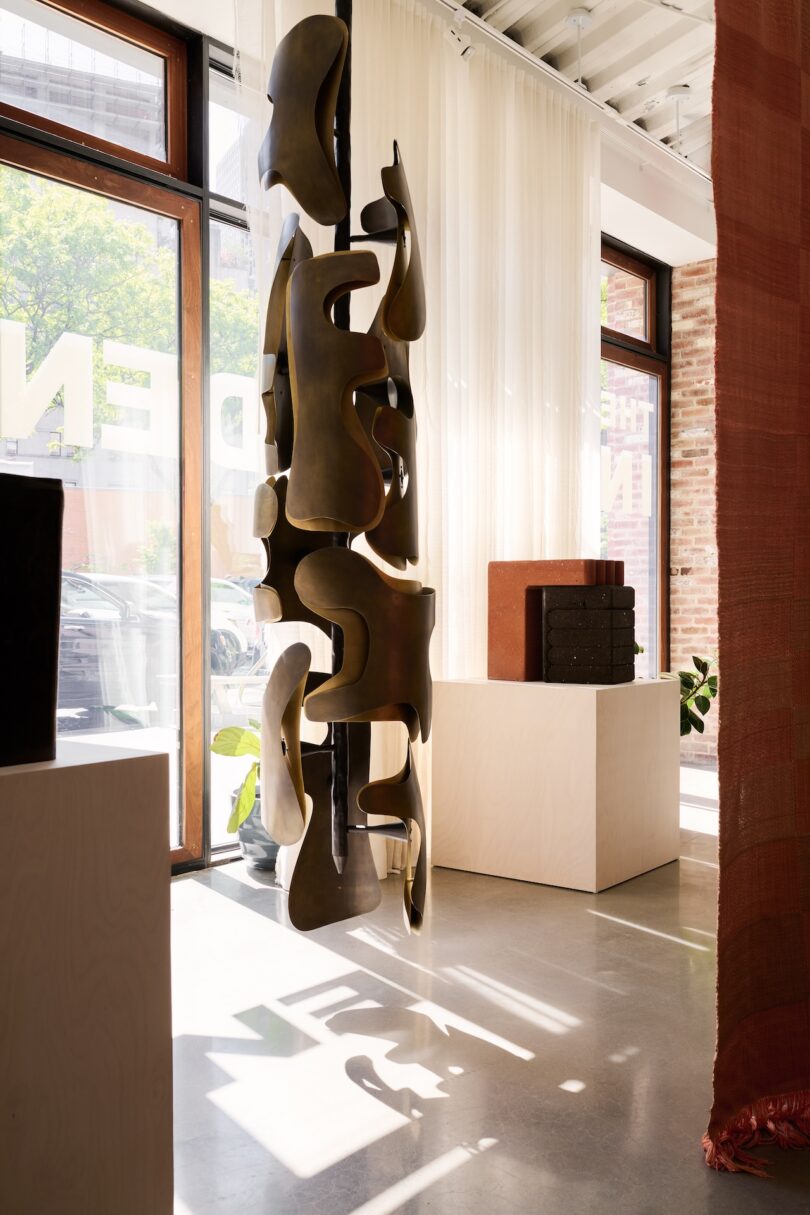 Abstract, bronze-colored metal sculpture hangs in a sunlit gallery space with large windows, light curtains, and pedestals displaying other art objects.