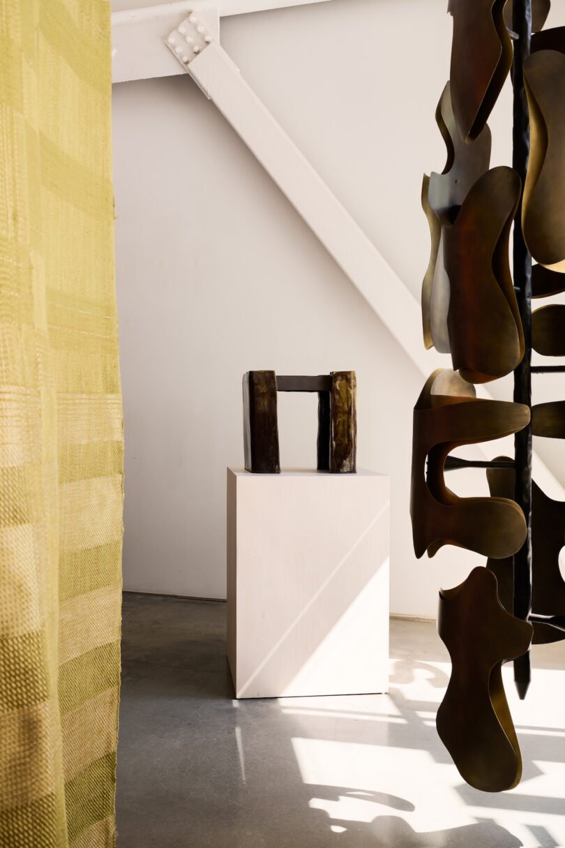 A small abstract sculpture sits on a white pedestal in a sunlit gallery space, with a yellow textile and a larger hanging metal sculpture partially visible.