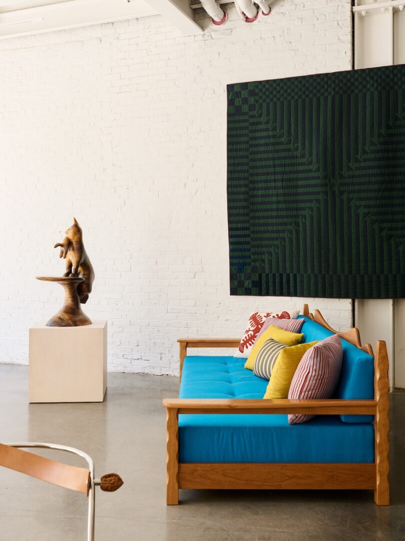 A modern living space with a wooden sofa featuring bright blue cushions and colorful pillows, a wooden bear sculpture on a pedestal, and a geometric dark green textile on a white brick wall.