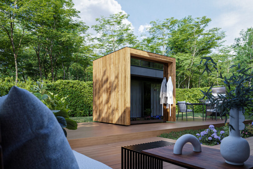 A small, modern wooden garden shed sits on a wooden deck surrounded by greenery, outdoor furniture, and flowering plants