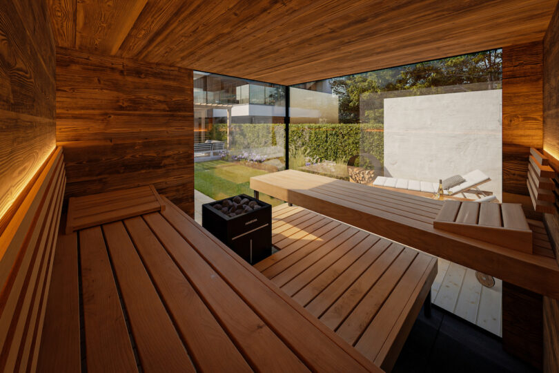 Modern wooden sauna interior with benches, a heater, and large glass windows showing a garden and outdoor lounge chairs