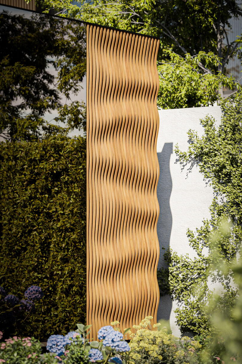 A tall, vertical wooden panel with a wavy pattern stands in a garden, surrounded by greenery, flowers, and a white wall