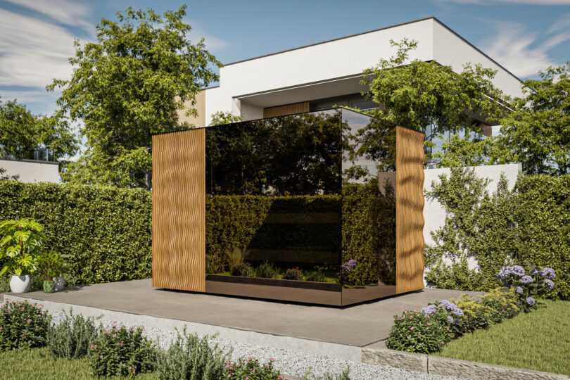 A modern garden office pod with Thermasol reflective glass walls and wooden paneling sits on a paved area surrounded by greenery and landscaping