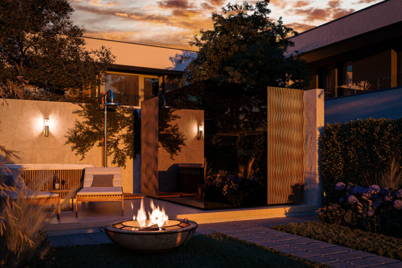A modern outdoor patio at sunset features a lit fire pit, a bench with cushions, wall-mounted lights, and surrounding trees and shrubs