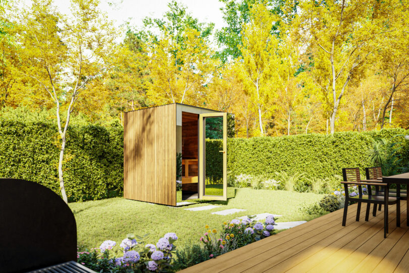 A small wooden shed with an open door sits in a sunny, fenced backyard with green grass, blooming flowers, and a patio featuring a Thermasol table and chairs