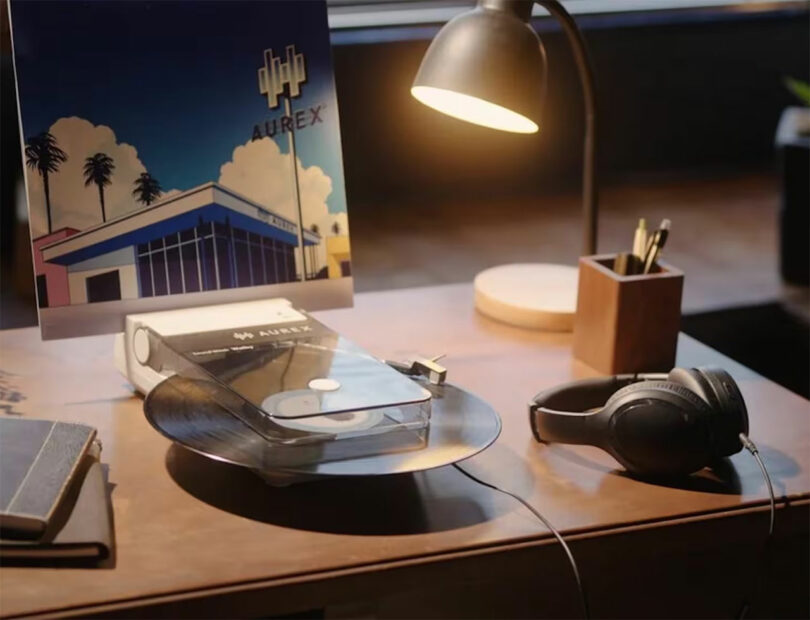 A modern transparent AX-RP10 record player spins a vinyl next to a pair of headphones, a desk lamp, stationery, a notebook, and a poster featuring a building and palm trees.