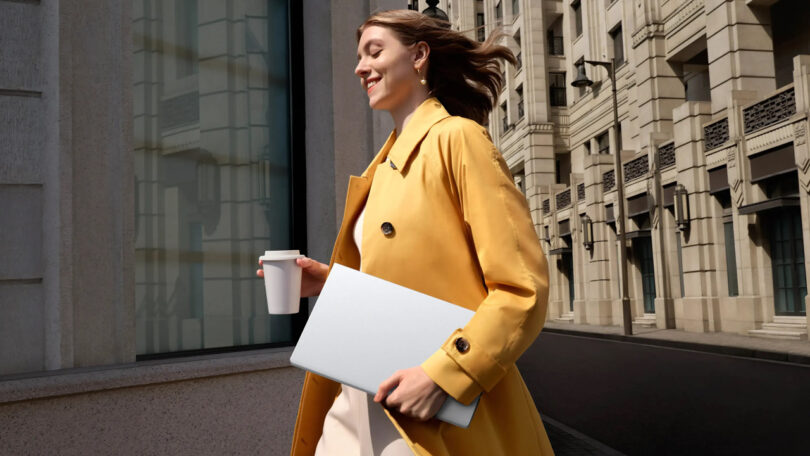Woman in a yellow coat walks outside, holding a coffee cup and a closed laptop, with city buildings in the background.