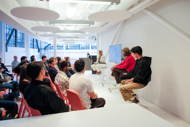 A group of people sits in an open, modern room listening to three speakers seated at the front near a large display screen