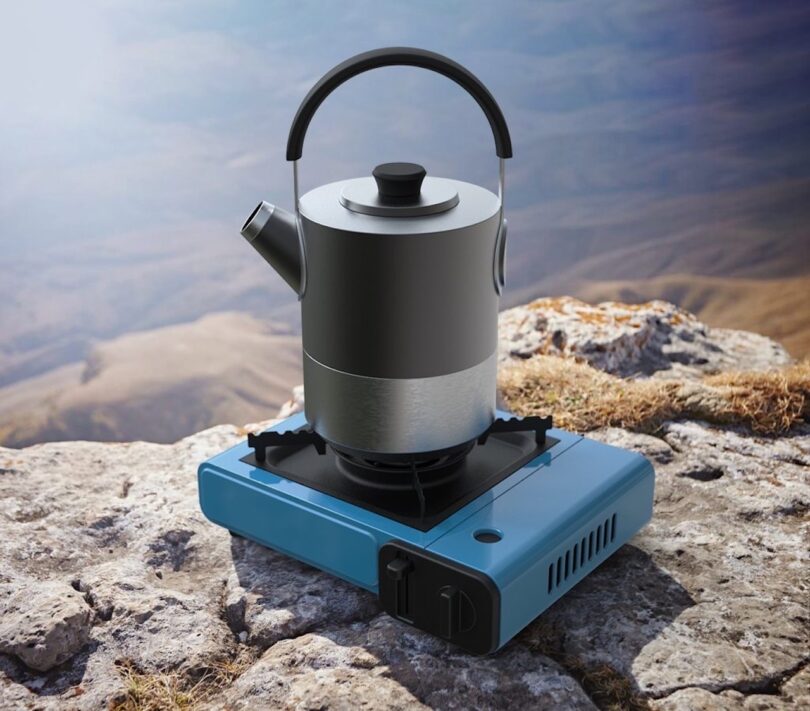 A metal kettle sits on a blue portable gas stove placed on a rocky surface outdoors with mountains in the background