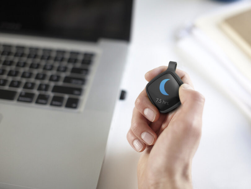A hand holds a fitness tracker device showing a crescent moon icon and "7.5 hrs" next to an open laptop