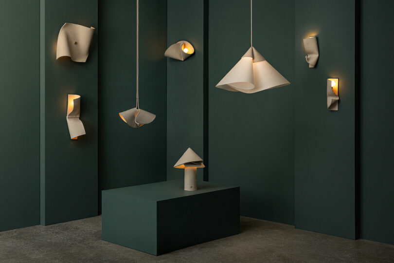 A set of modern sculptural lamps by Virginia Sin lighting, with soft, folded white shades, is displayed against dark green paneled walls and a matching pedestal in a minimalist gallery setting