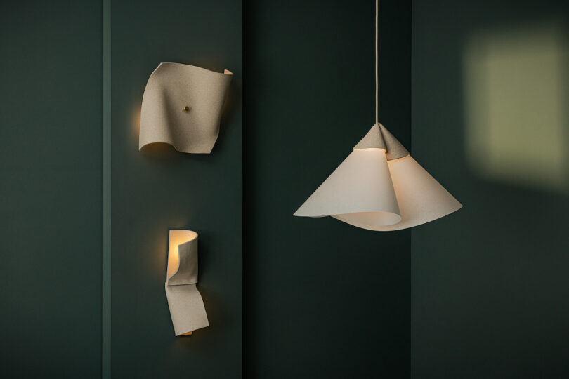 Two wall-mounted lights and one pendant light with folded, sculptural white shades from Virginia Sin lighting are set against a dark green background