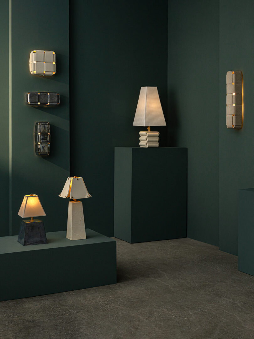 A display of modern lamps and Virignia Sin lighting wall sconces with geometric shapes, set against dark green walls and platforms in a minimalist gallery space